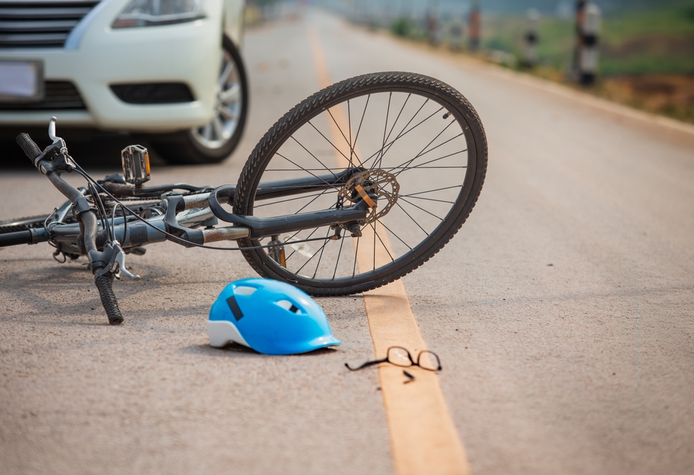 Bicycle accident scene showing a fallen bike, helmet, and broken glasses near a white car on the road.