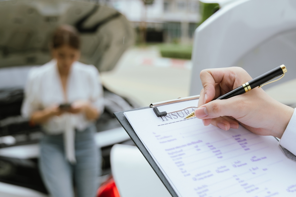 Insurance paperwork being filled out with a damaged car and driver blurred behind.