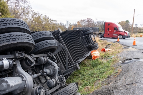 Overturned semi-truck lying on its side off a rural highway with debris and traffic cones nearby.