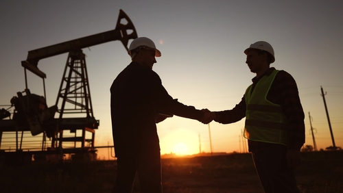 Two oil workers shaking hands near a pump jack during sunset.