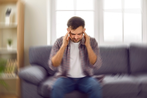 A man sitting on a couch holding his head in pain, showing symptoms of a delayed brain injury after an accident.